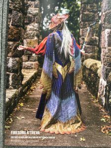 Lazzaro Cosplay's photo shows the sweeping train at the back of the outer robe with its layers of dark blue, pale silver/blue and gold feathers. The mask is seen in profile with long silver hair cascading from the back of the mask. The setting is an ancient stone causeway between high walls with fallen leaves lying on the ground.
