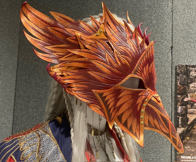 The mask of Firesong K'Treva by Patterner Cosplay. Layers of leathery segments evoke armour, flames and feathers at one, coloured in tones of gold and bronze. The mask has a long beak and is shaped like a bird of prey's head.
