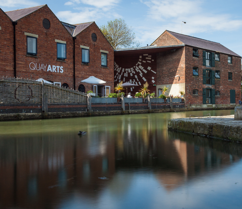 Quay Arts Centre is on the quay of the old port of Newport, on the River Medina. Here the old red brick buildings are reflected in the sunlit blue water of the river.