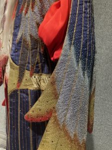Detail of outer robe sleeves. The robe is patterned in bands of dark blue/green, silver and gold edged with reddish bronze and textured with tiny light-reflecting gems. The sleeves of the scarlet under-robe are visible.