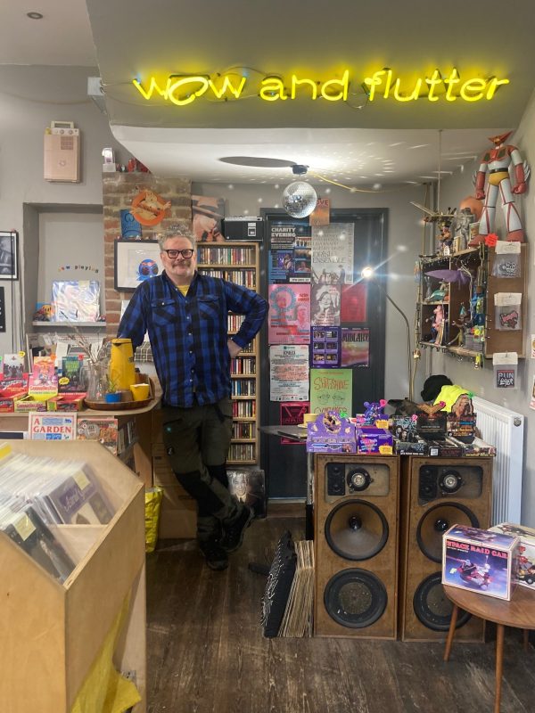 The interior of Wow and Flutter with Gareth smiling under the bright yellow neon sign showing the shop's name. Gareth is tall, with grey hair, wearing a dark blue open-necked shirt and dark trousers. The shop is crammed with art, vinyl, zines, comics and Japanese toys. Photo by Helen McCarthy