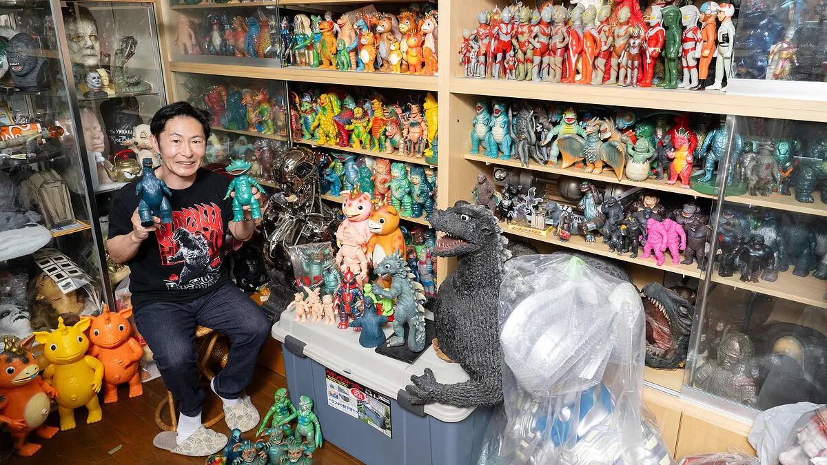 Veteran kaiju collector Nabe Yakan sits in front of shelves lined with some of his treasures - colourful and now highly valuable plastic toys depicting kaiju and tokusatsu characters. Nabe wears black trousers, grey slippers and a black and white T-Shirt with the logo GODZILLA in bright red behind an image of the King of the Monsters, and holds a toy in each hand.
