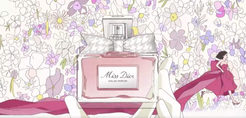 A frame from the new Dior US ad for Miss Dior perfume. A gamine girl in an unravelling deep pink ballgown sprints across a background of flowers behind a huge gloved hand holding a soft pink bottle of Miss Dior perfume.
