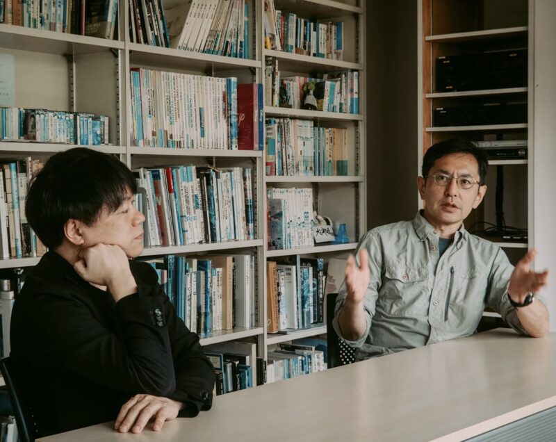 Kenichi Yoda (black top) and Goro Miyazaki (blue-grey shirt) seated behind a table in a book-lined conference room at Studio Ghibli. Photo ©Kenichi Takahashi